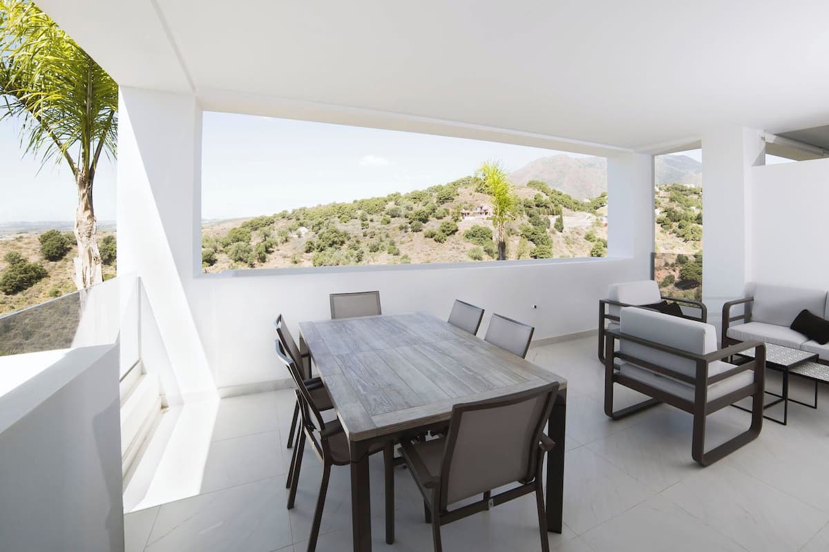 Un espace repas extérieur moderne, doté d’une grande table et de chaises, d’un canapé et offrant une vue sur un paysage vallonné.