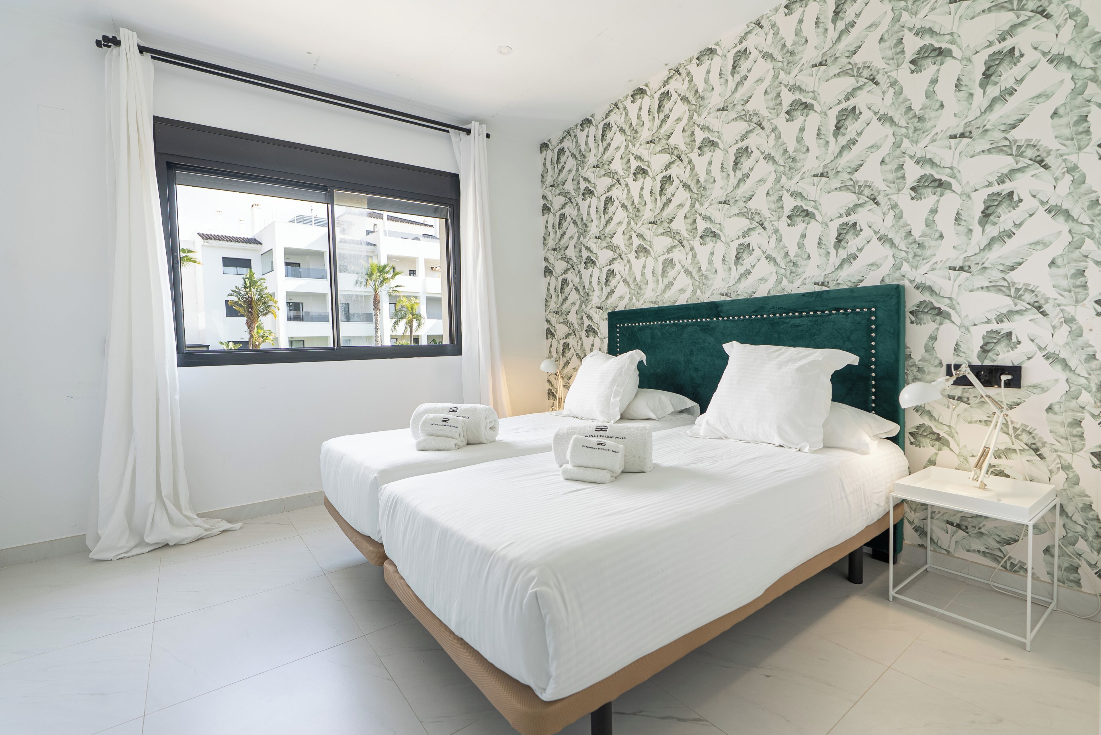 A bedroom with two beds, a window with curtains, a green headboard, and a patterned wallpaper.