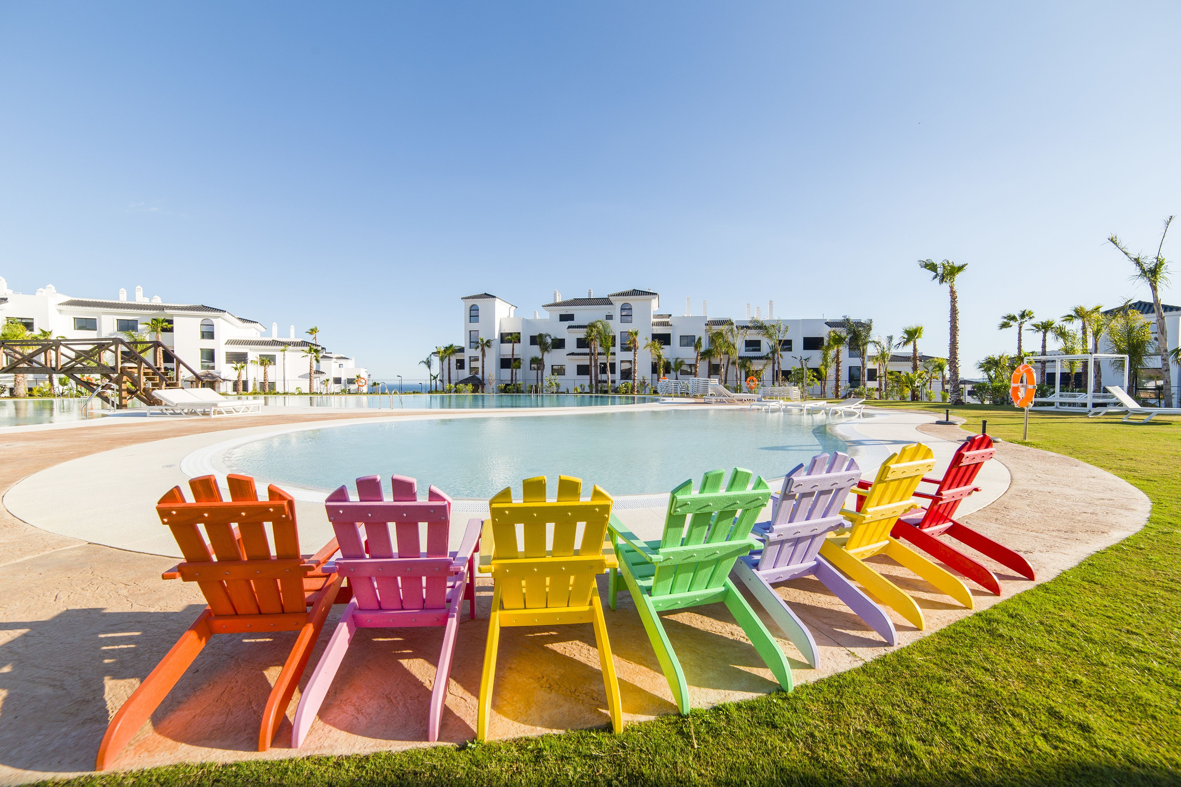 A poolside area with colorful Adirondack chairs, a clear blue sky, and modern buildings in the background.