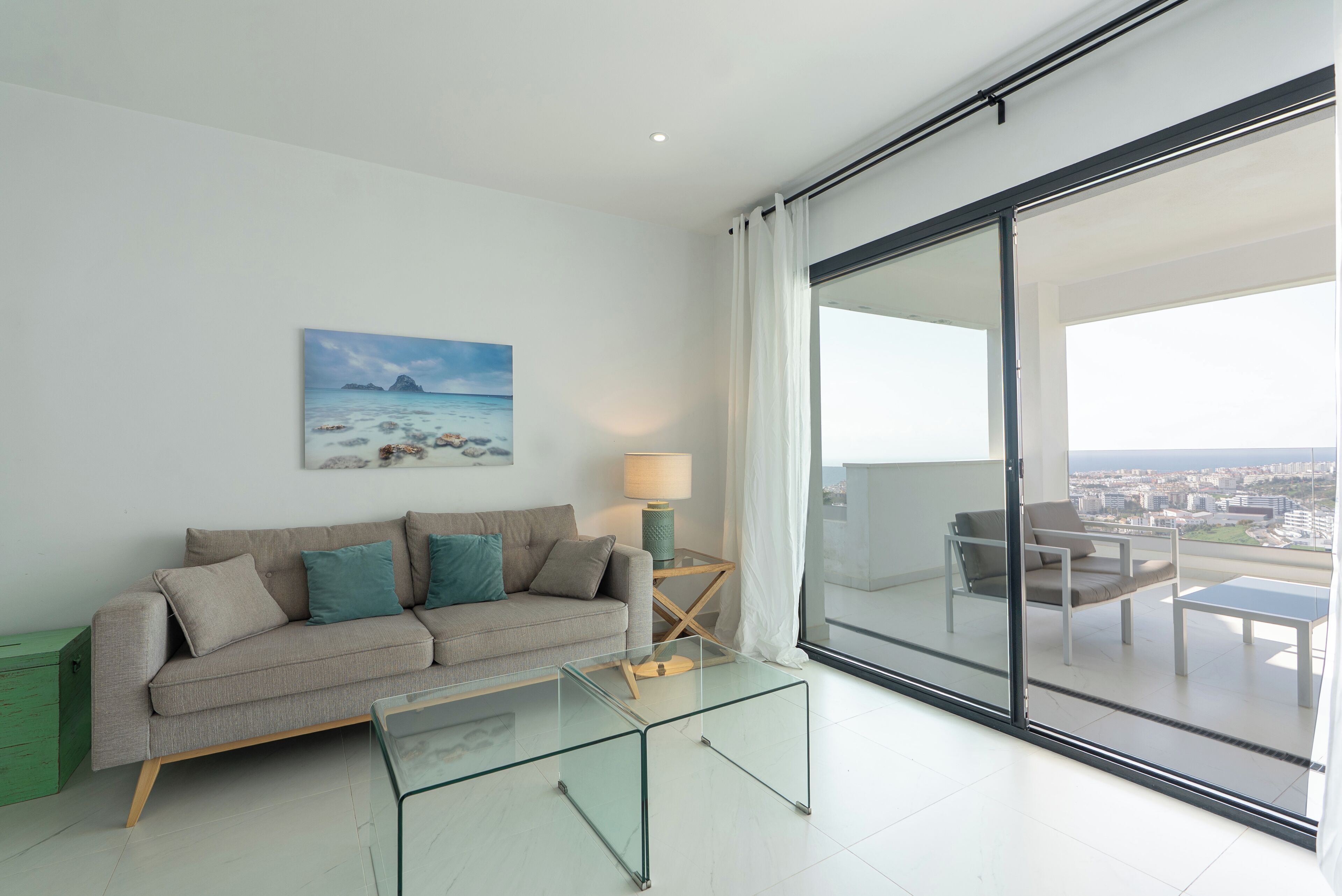 Een moderne woonkamer met een glazen salontafel, een bank met kussens, een schilderij van een strandlandschap en uitzicht door een schuifdeur op een balkon met een tafel en stoelen.