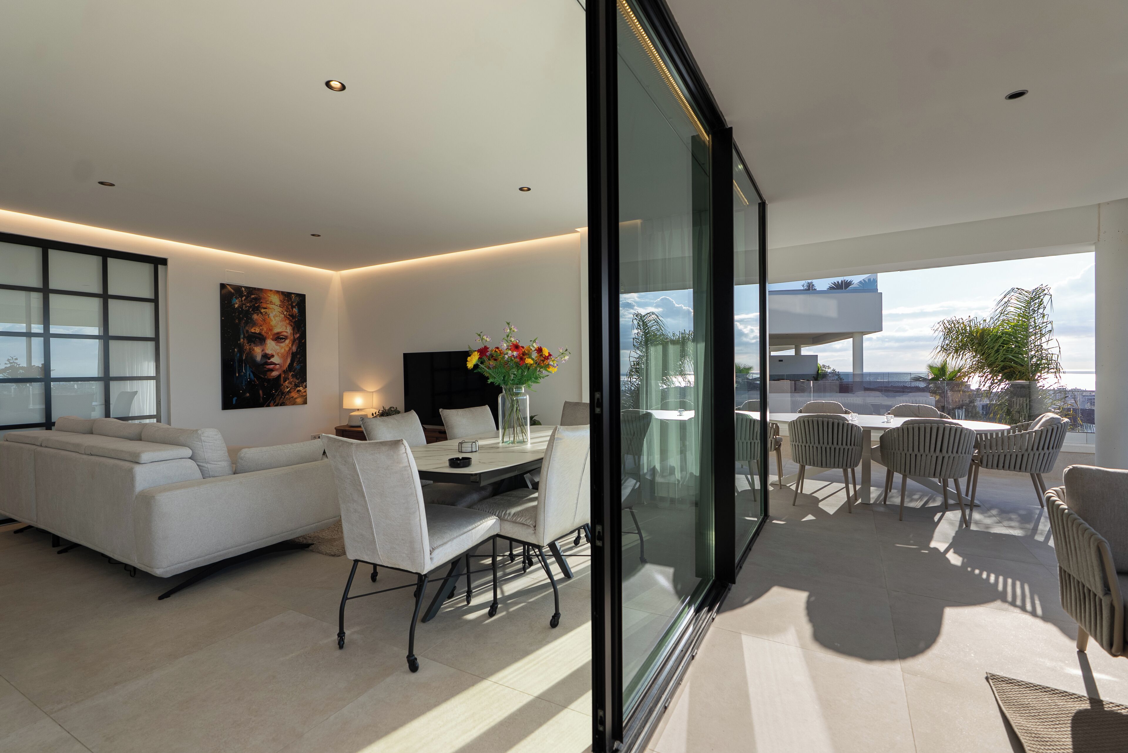 A modern living room with a sofa, dining table, and chairs, a large window, and a balcony with outdoor seating.