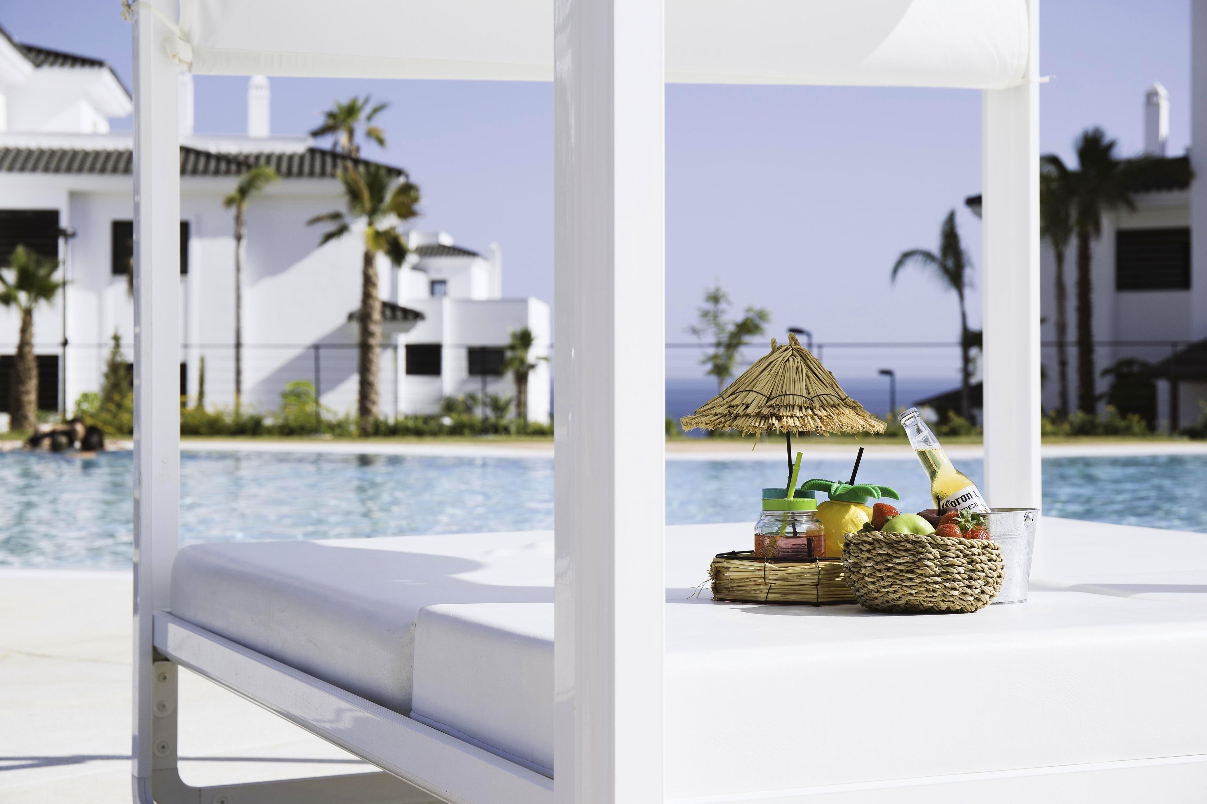 Escena junto a la piscina con una sombrilla blanca, una botella de cerveza Corona y una cesta de frutas.