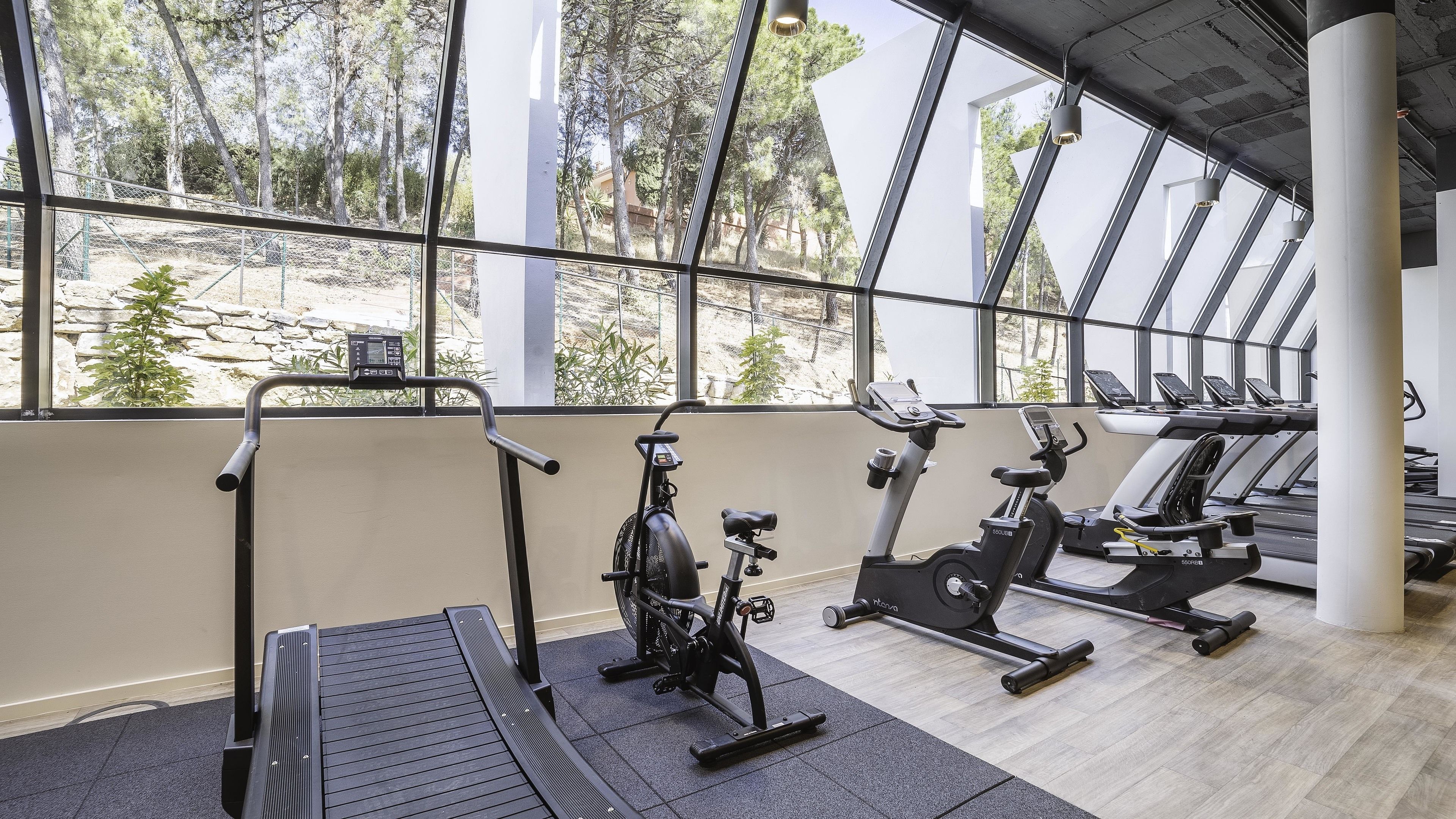 Un gimnasio con equipamiento de ejercicio, grandes ventanales y vistas a árboles.