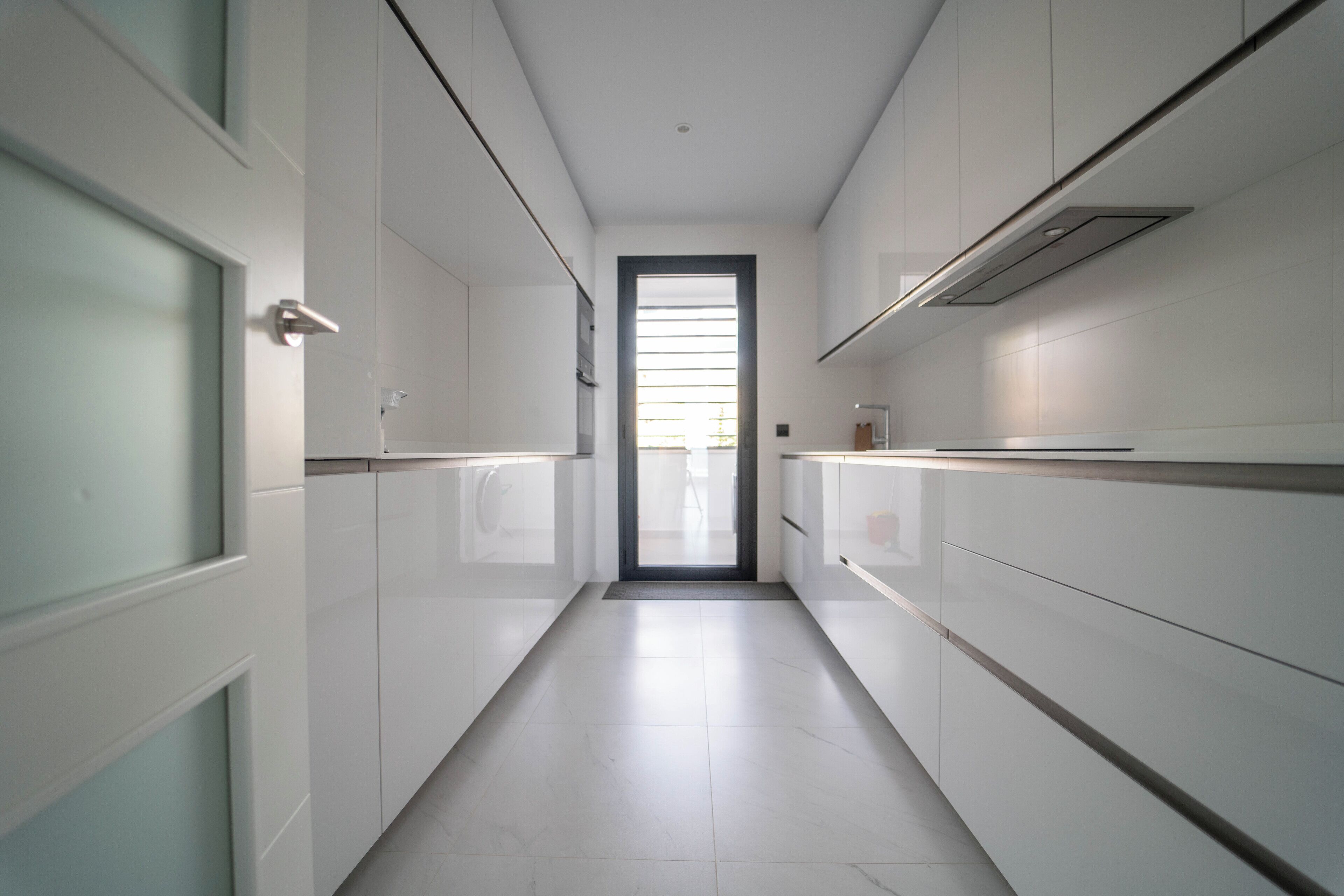 A narrow, modern kitchen with white cabinetry, a visible door, and a window at the end of the corridor.