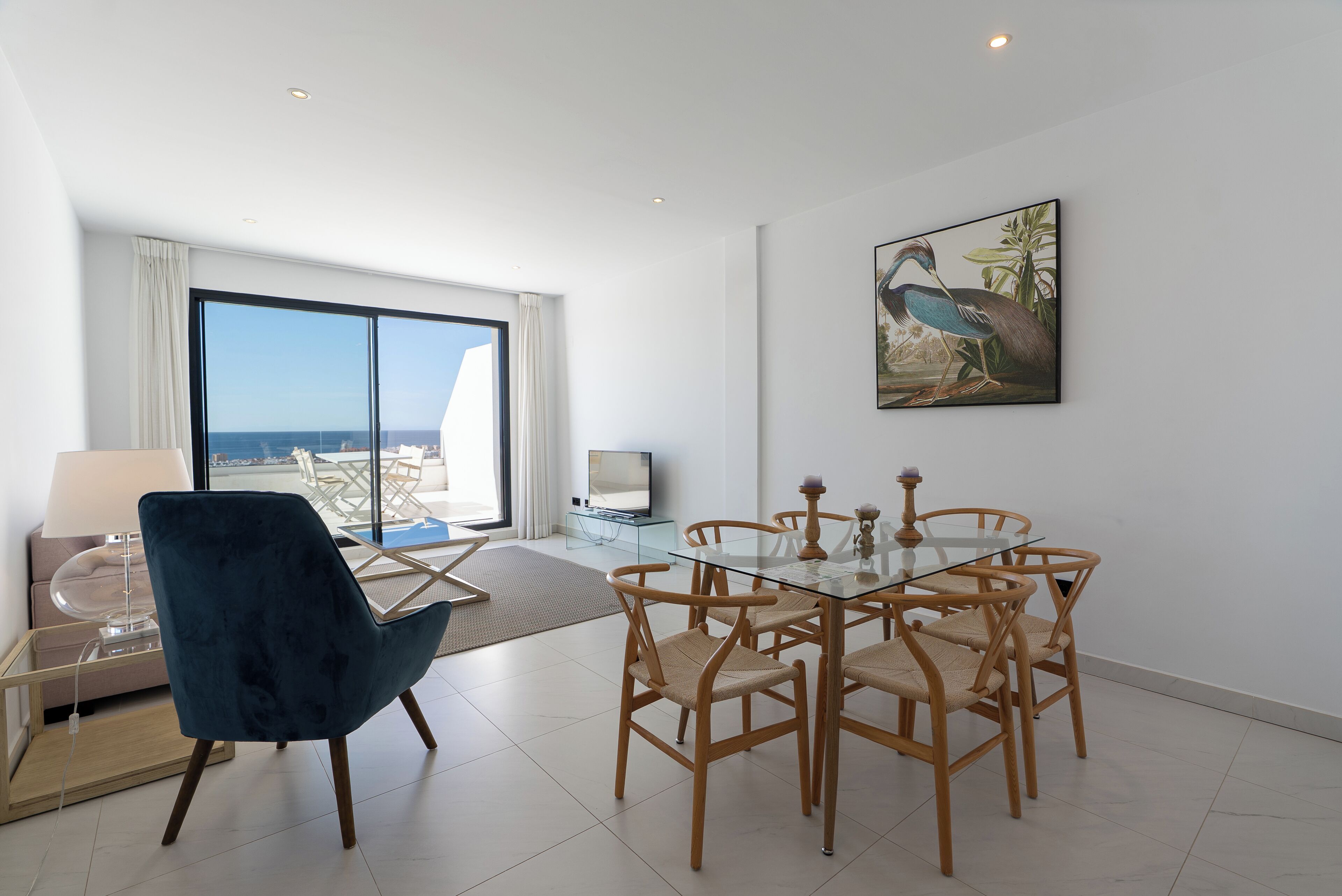A modern dining area with a glass table and wooden chairs, a blue chair, and a painting on the wall.