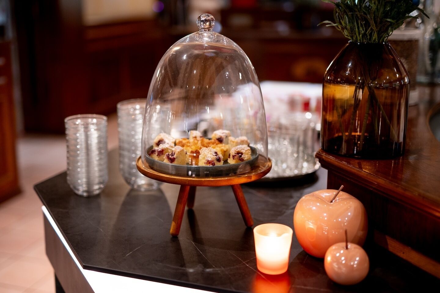 Une présentation de desserts sous un cloche de verre, agrémentée de bougies et d’un vase rempli de verdure, le tout posé sur une table en bois.