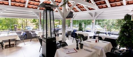 An outdoor dining area with tables and chairs, a grill, and a view of the surrounding greenery.