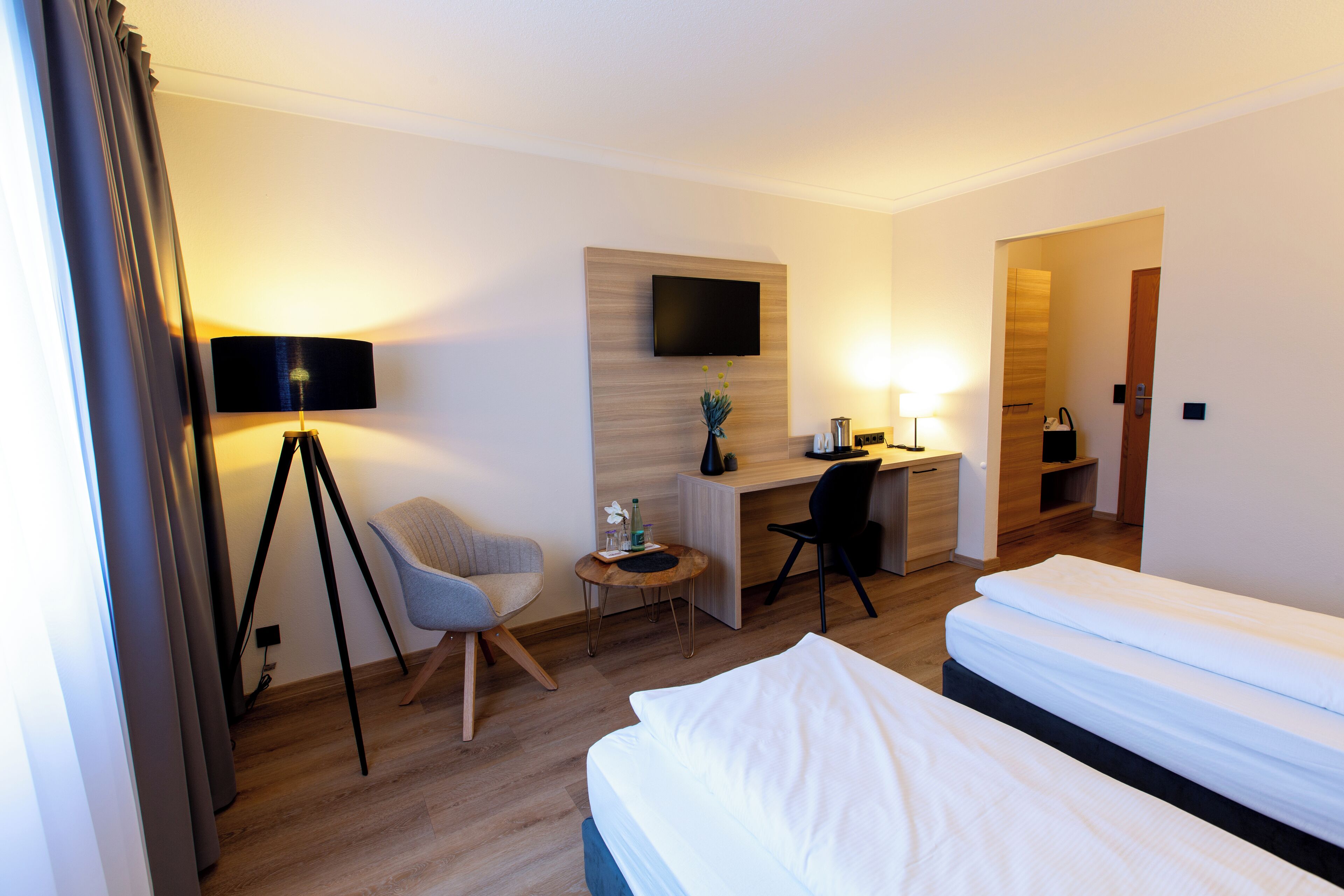 Quarto de hotel com cama, escrivaninha, cadeira e televisão fixada na parede.