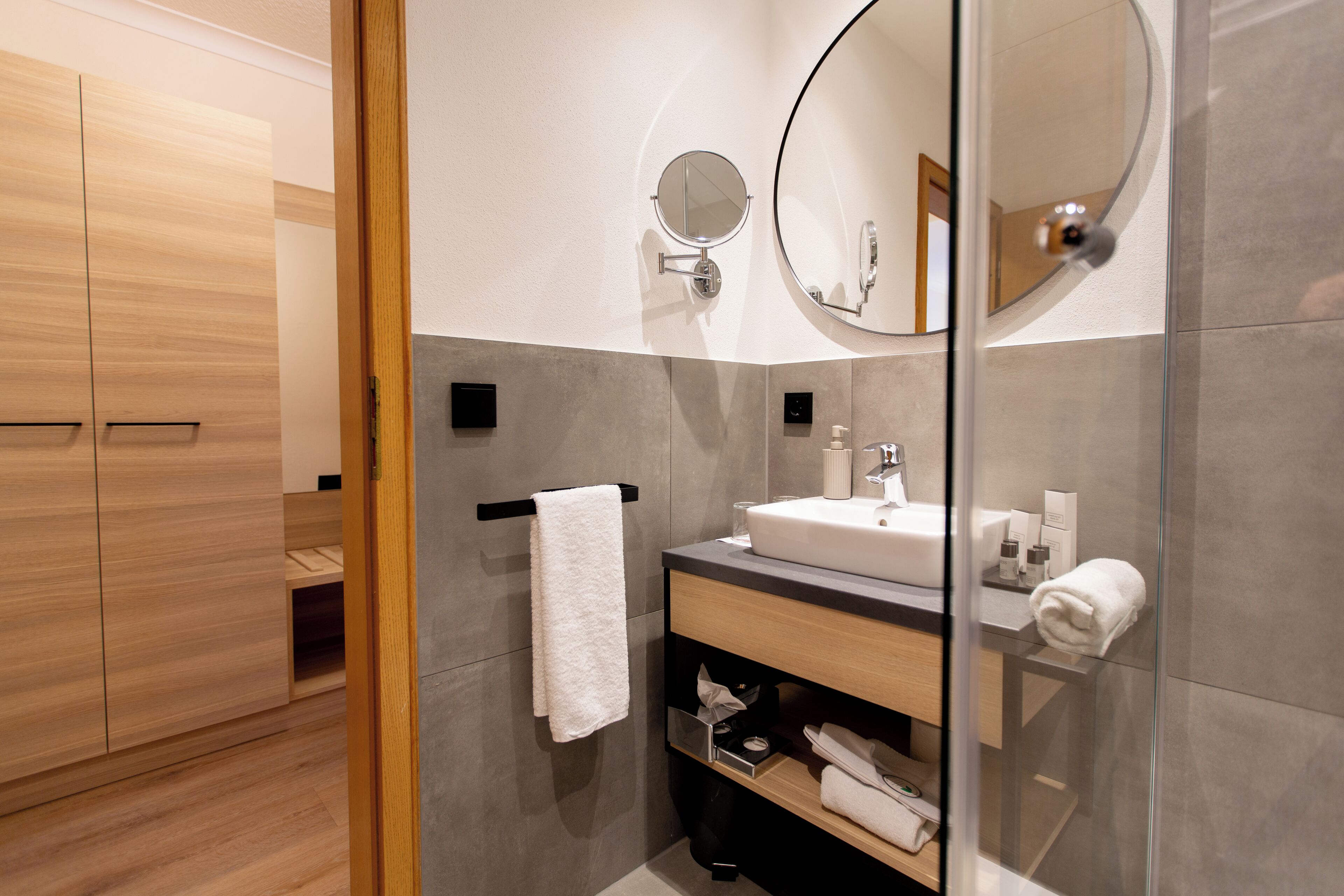 Un baño moderno con un armario de madera, cabina de ducha de vidrio, lavamanos blanco y espejo redondo.