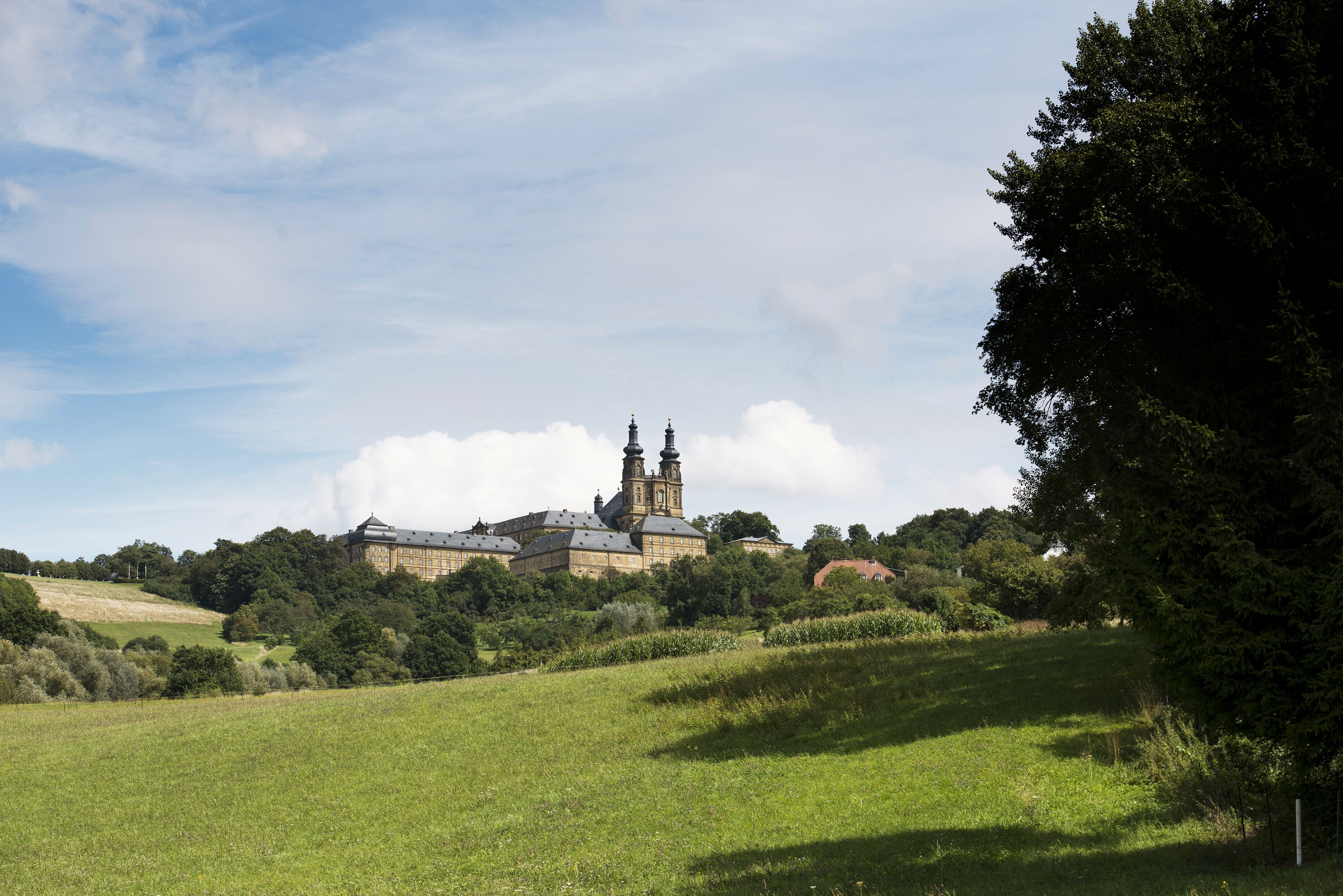 A large, historic building with two towers, surrounded by lush greenery and fields.