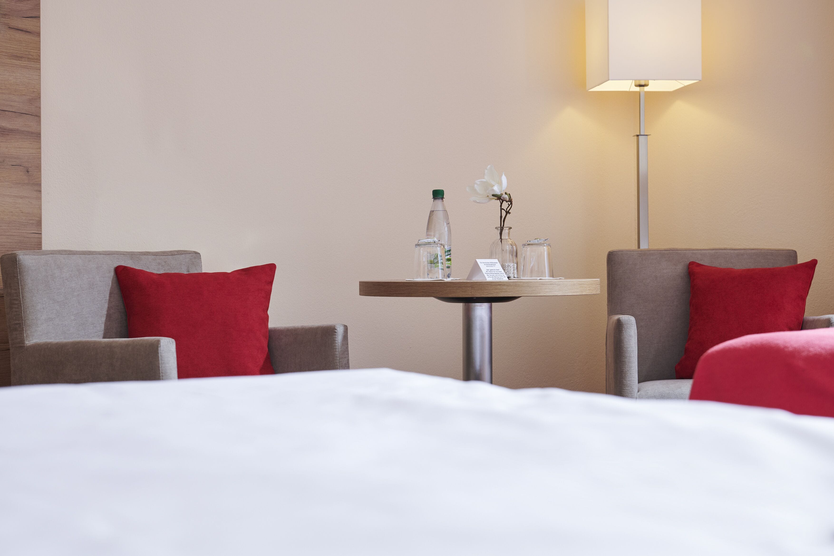 Habitación de hotel con dos sillones, una mesita redonda con lámpara y una cama con almohadas rojas.