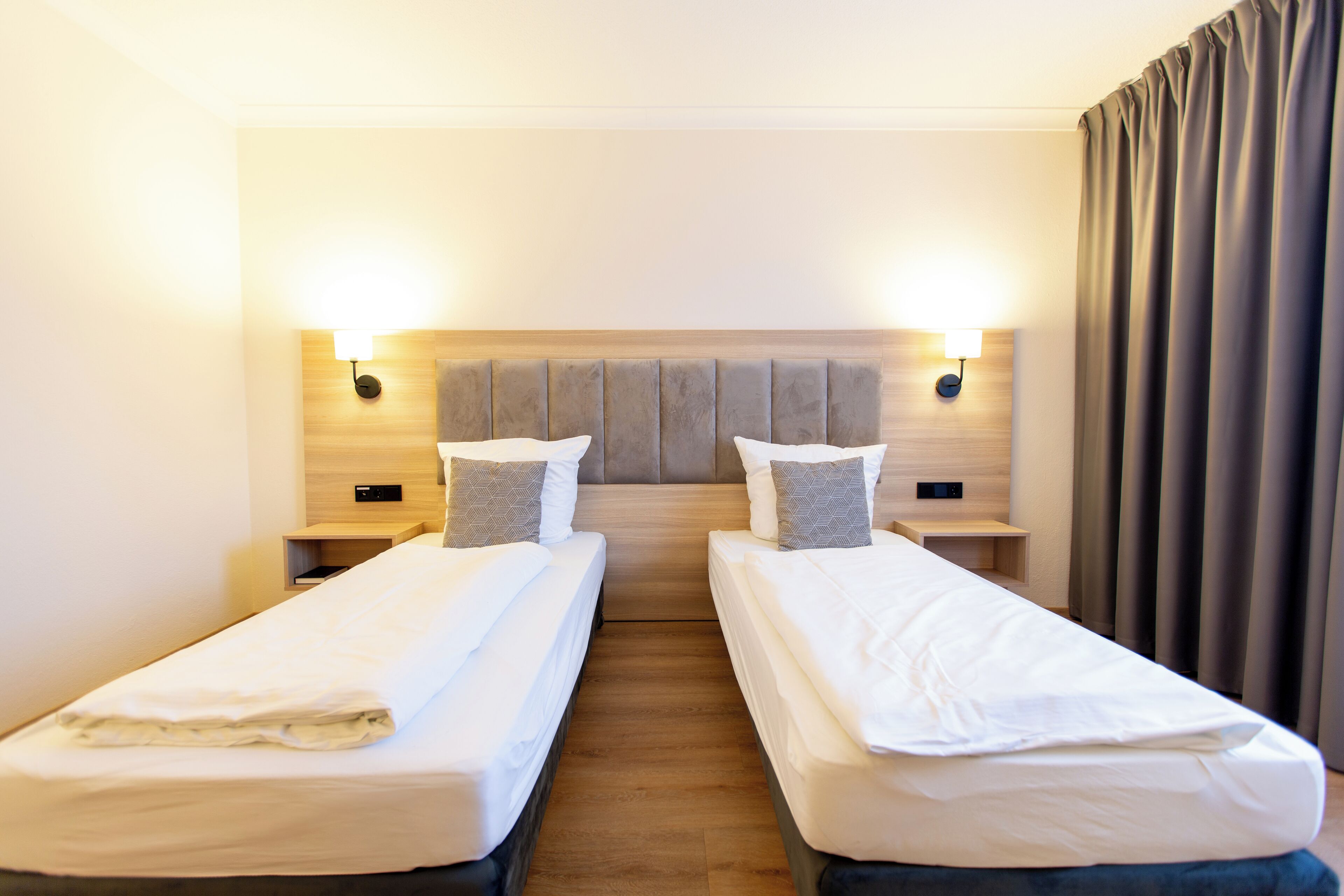 A hotel room with two single beds, a wooden headboard, bedside lamps, and a curtain.