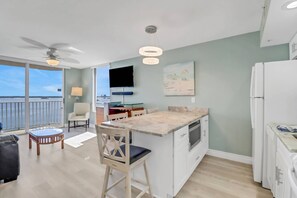 A modern kitchen with a marble countertop, a dining area with a table and chairs, a balcony with a view of the water, and a ceiling fan.