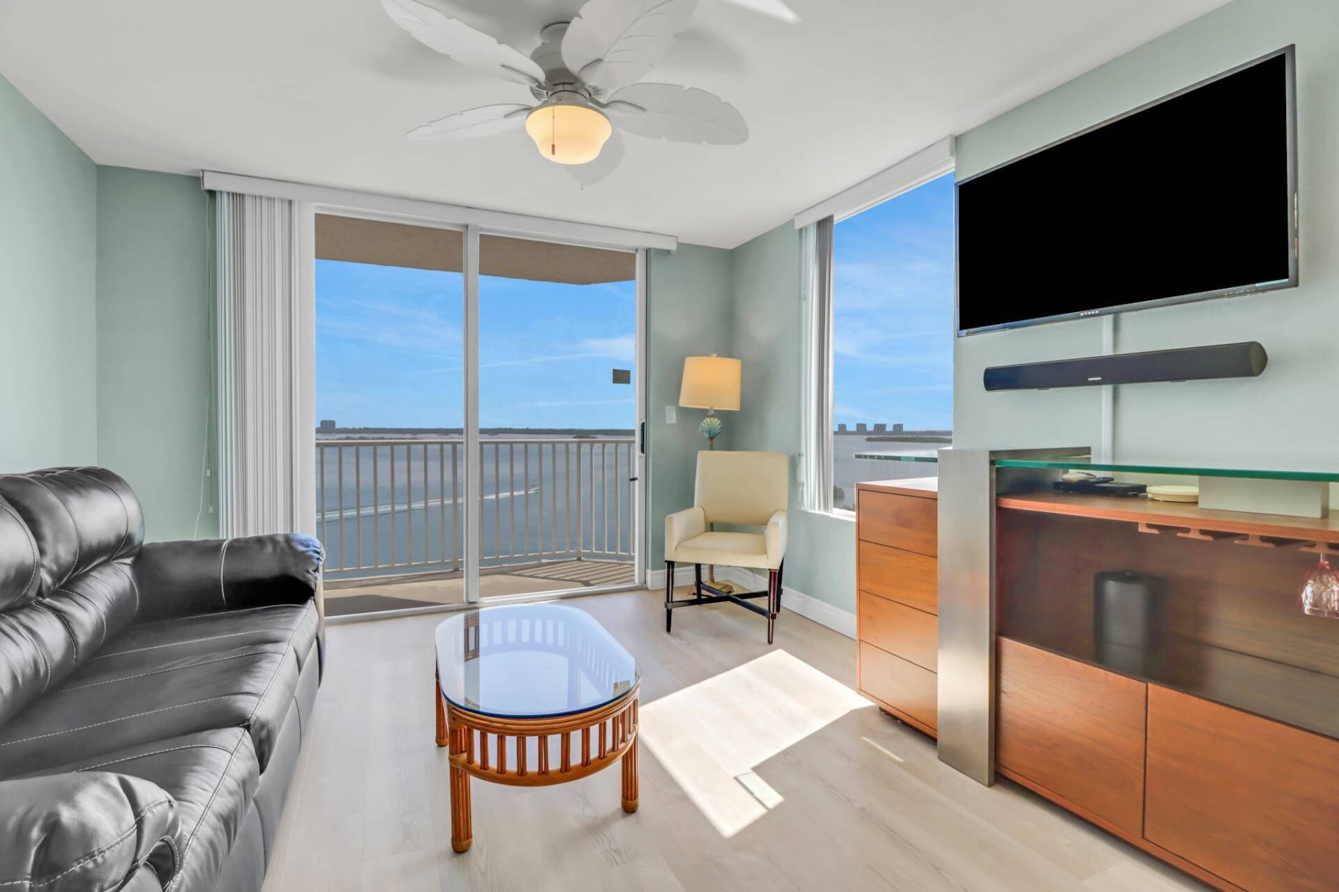 Una habitación de hotel moderna con balcón, un sofá de cuero, una mesa de centro de vidrio, un mueble de madera y un televisor de pantalla plana.