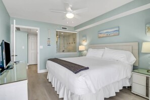 A bedroom with a large bed, a ceiling fan, a window with blinds, a bedside table with a lamp, and a door leading to another room.