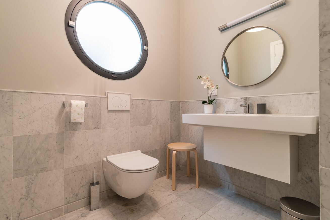 A modern bathroom with a toilet, a small stool, a round mirror, and a sink.