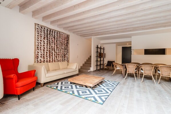 Una sala moderna con un sillón rojo, sofá beige, mesa de centro de madera, alfombra estampada y un comedor con sillas de mimbre.