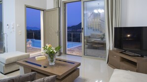 Ein modernes Wohnzimmer mit Flachbildfernseher, Couchtisch mit Vase und Sofa. Ein Glastür öffnet sich zu einem Balkon mit Pool und Bergblick.