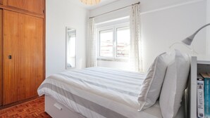 A bedroom with a bed, a wooden wardrobe, a window with curtains, a hanging light fixture, and a bookshelf with books. - Experience relaxing holidays (Estoril)