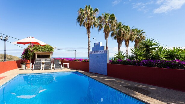 Una piscina con sillas de descanso y una sombrilla, rodeada de palmeras y flores púrpuras.