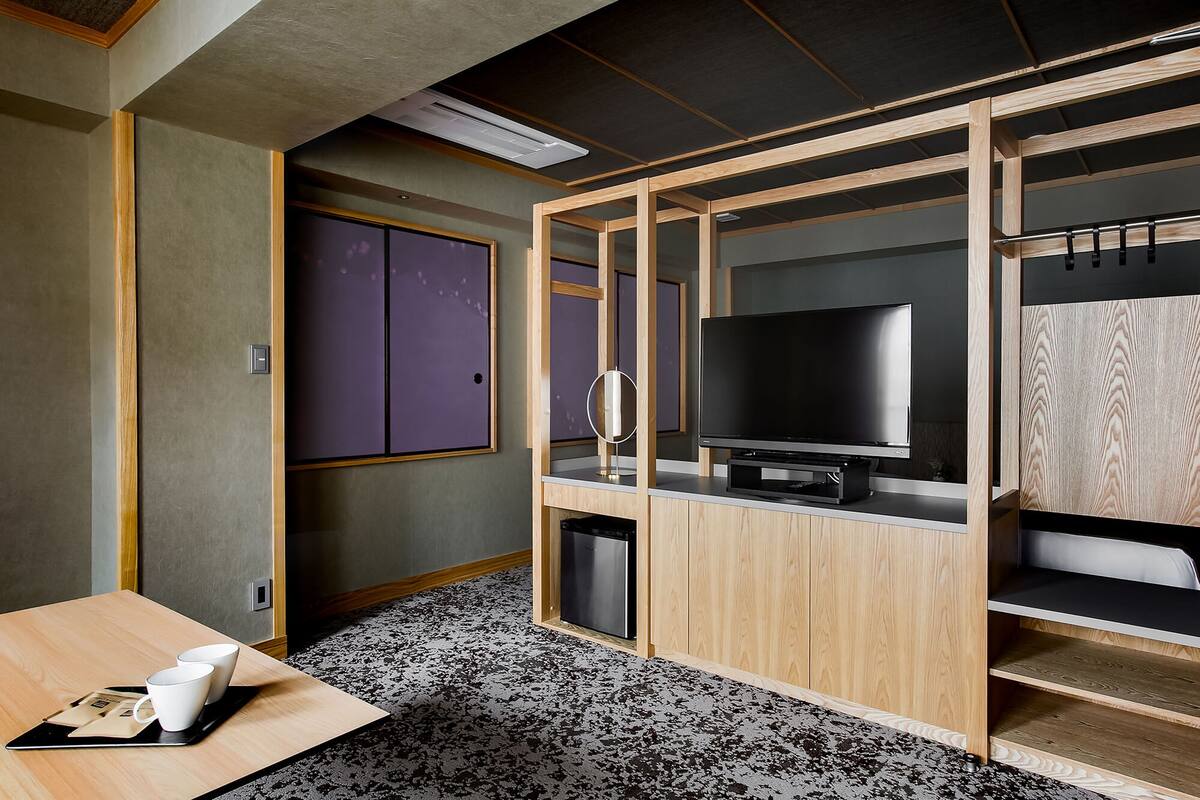 A compact hotel room with a wooden TV stand, a small seating area with a table, and a compact wardrobe.