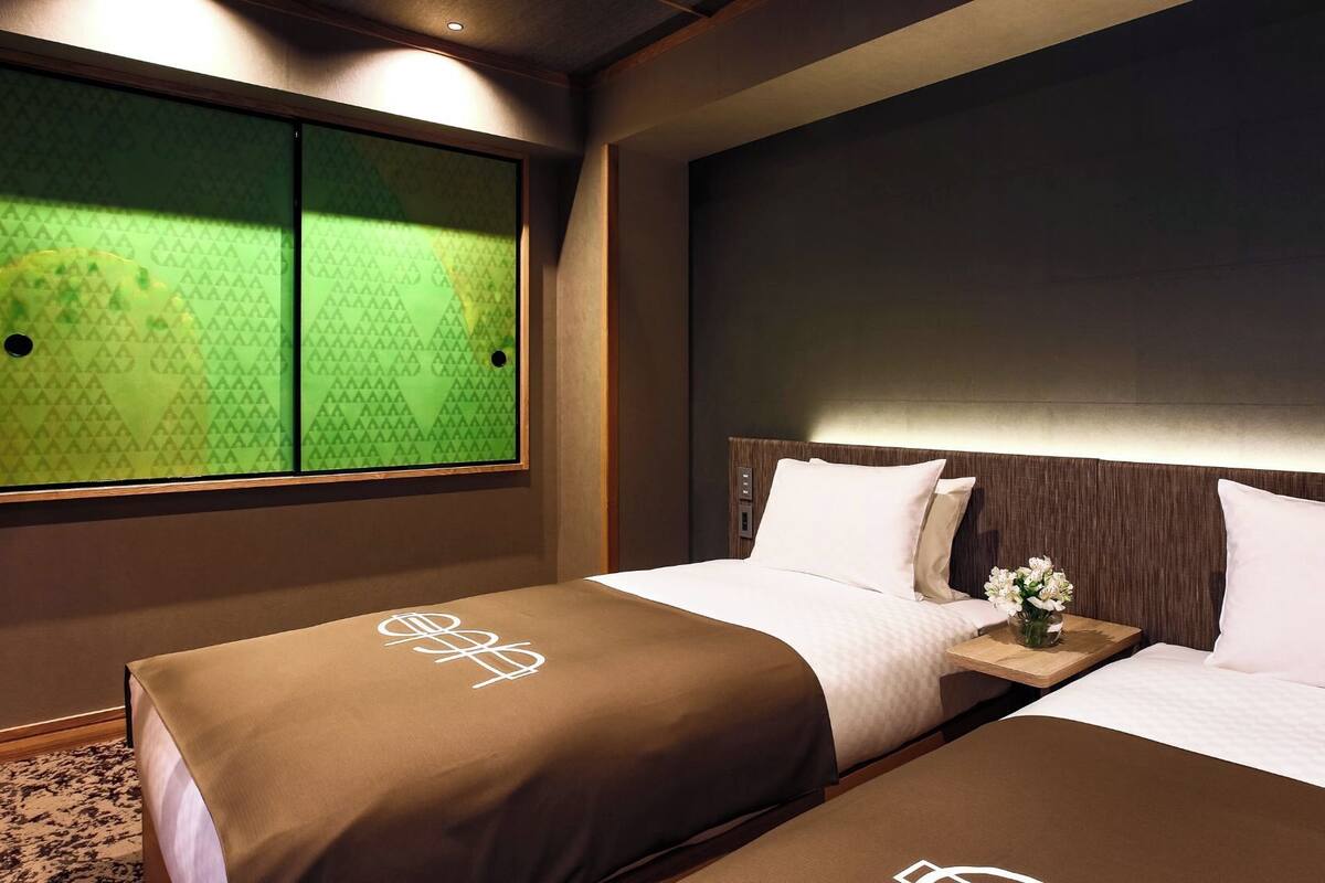 A hotel room with a bed, a nightstand, a vase with flowers, and a large window with a green patterned view.