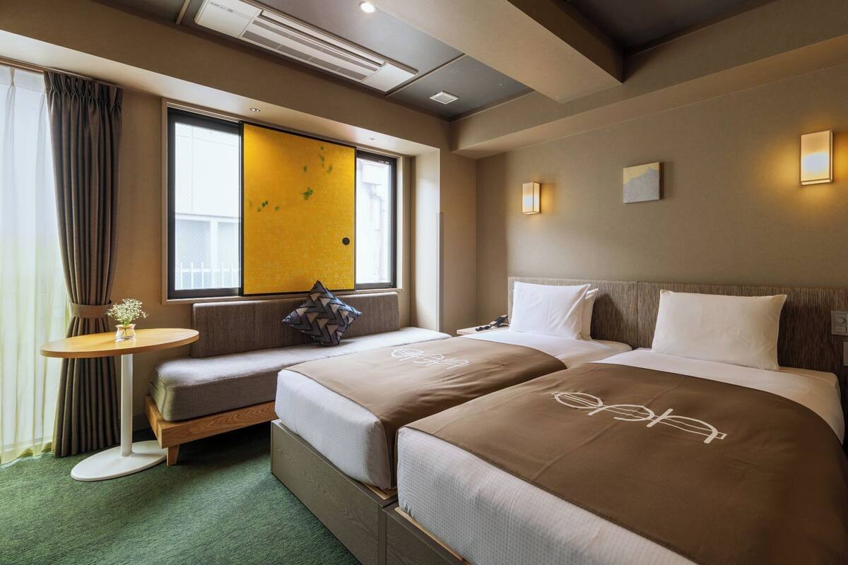 A hotel room with two beds, a small table, a window with curtains, and a wall-mounted light.