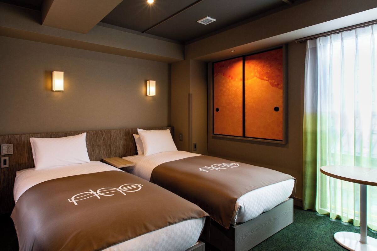 A hotel room with two beds, a small table, a window with curtains, and a wall-mounted artwork.