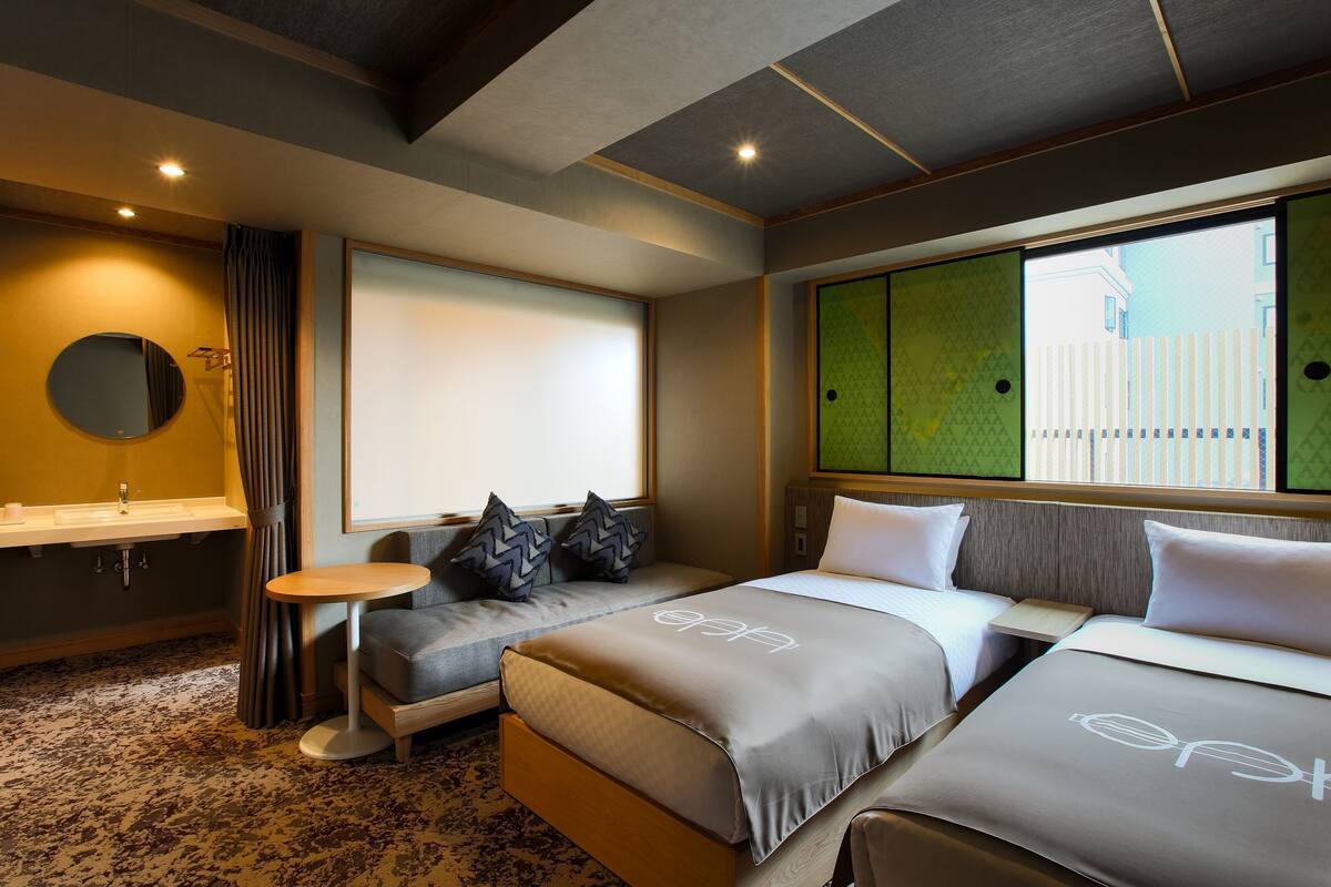 A hotel room with a sofa bed, a small round table, a sink, and a window with a view of a building.