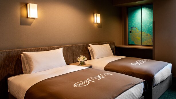 A hotel room with two beds, each with a brown bedspread and a white pillow. There are wall-mounted lights on either side of the beds, and a window with a view of the cityscape. - eph TAKAYAMA (Takayama)