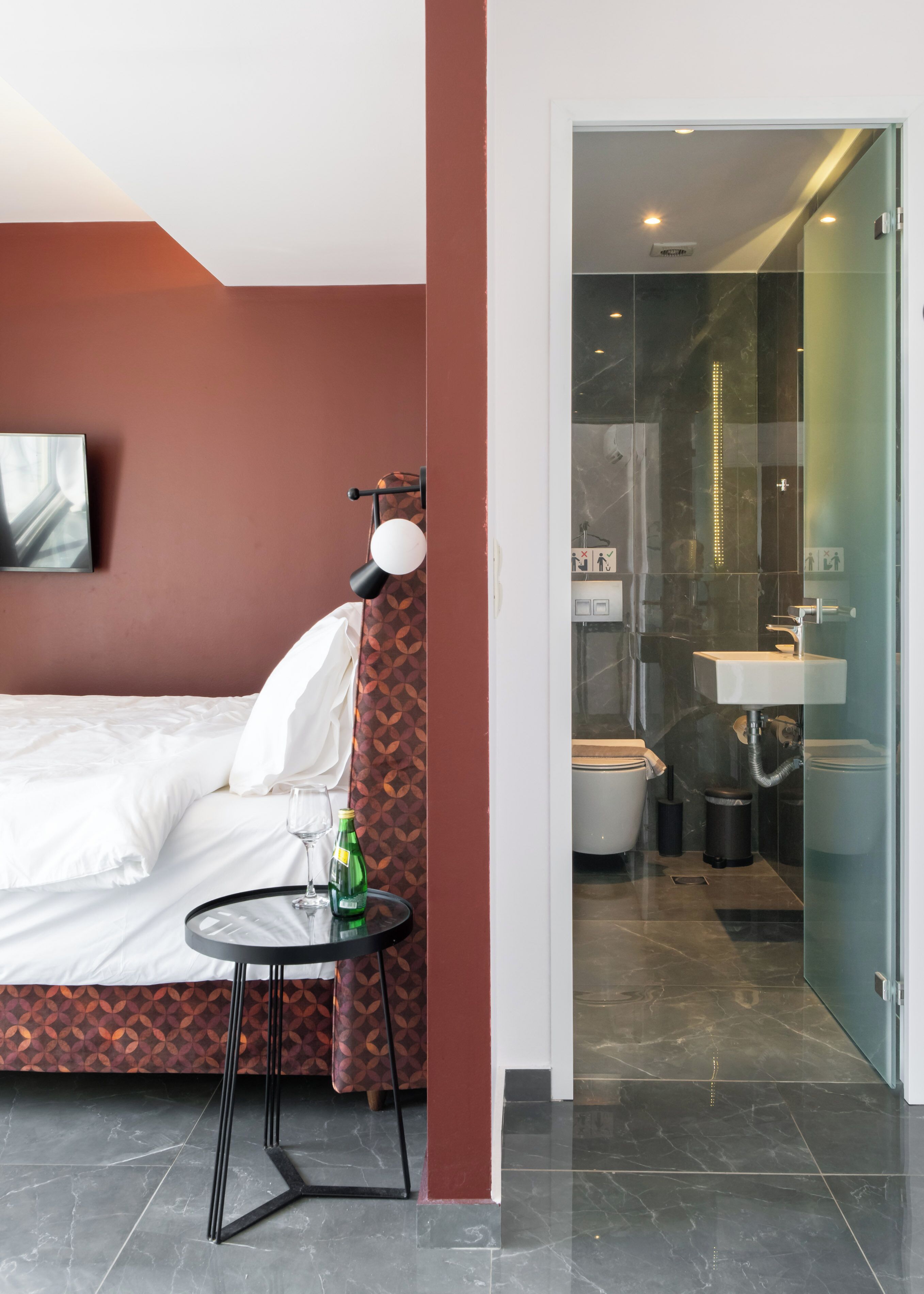 A modern hotel room with a bed, a small table with a glass, and a bathroom visible through a glass door.