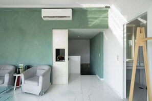A modern room with a white air conditioner, a white cabinet with a small window, two grey armchairs, a glass table, and a wooden ladder.
