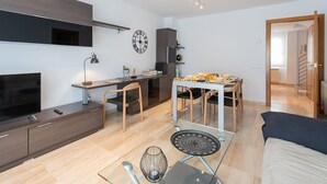 A modern living room with a sofa, a glass coffee table, and a TV. There is a dining area with a table and chairs, and a kitchen area with cabinets and a clock on the wall. - Duplex Ègara by Petit Luxe (Terrassa)