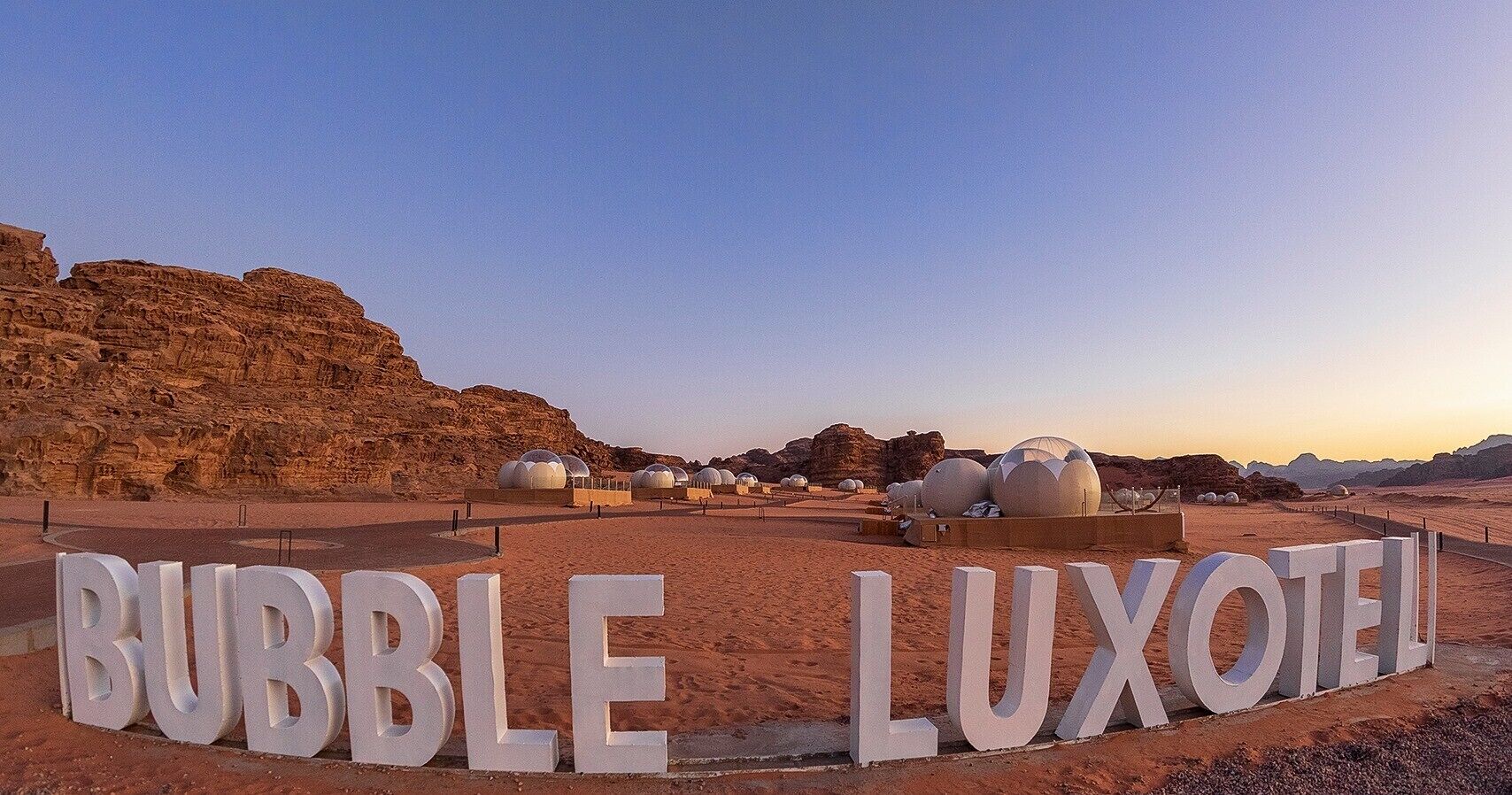Paisaje desértico con letras blancas grandes que deletrean "BUBBLE LUXOTEL" y varias estructuras en forma de cúpula.