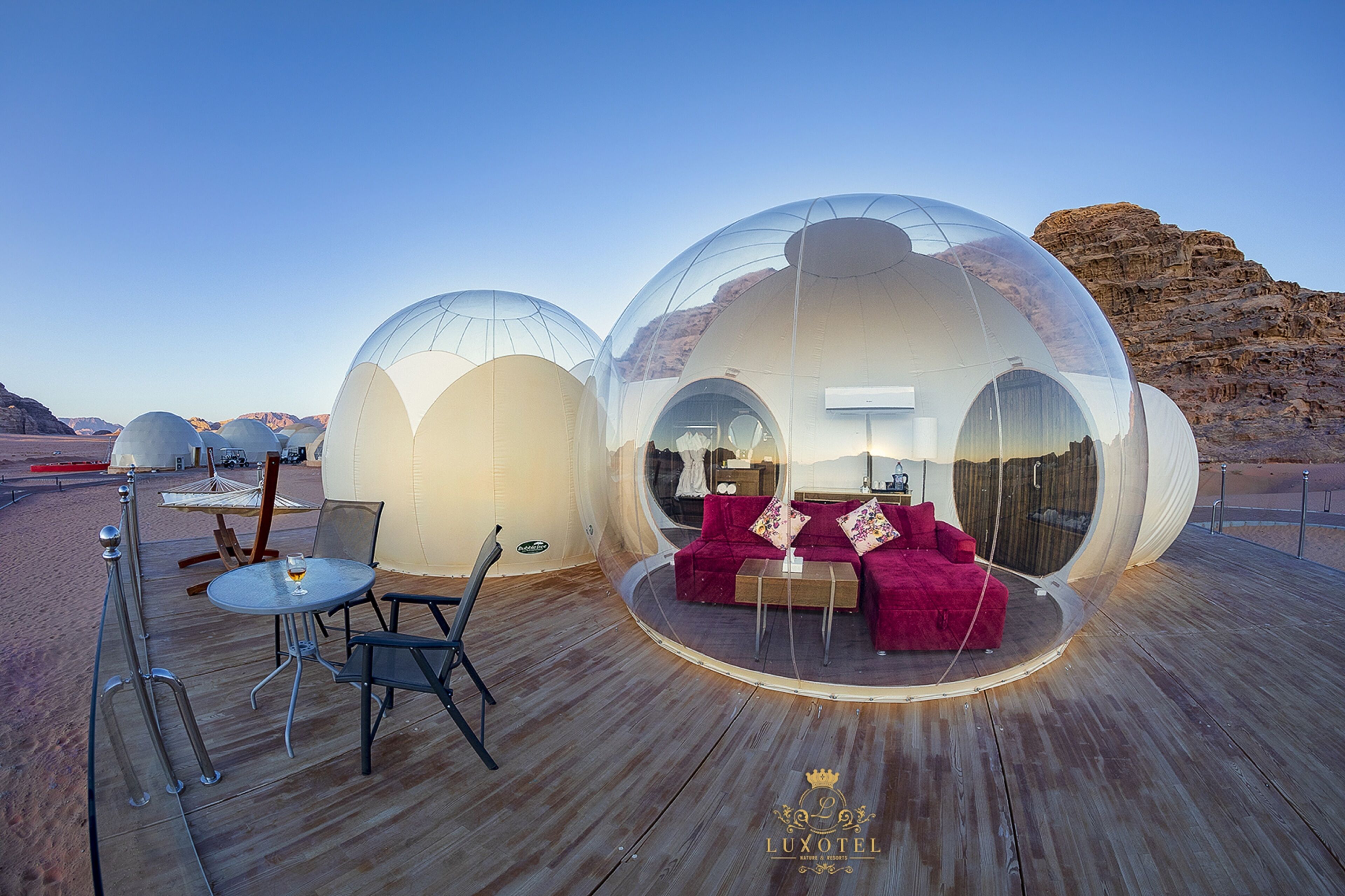 Transparent dome-shaped accommodations with a wooden deck, furnished with a sofa and chairs, and a table with a glass.