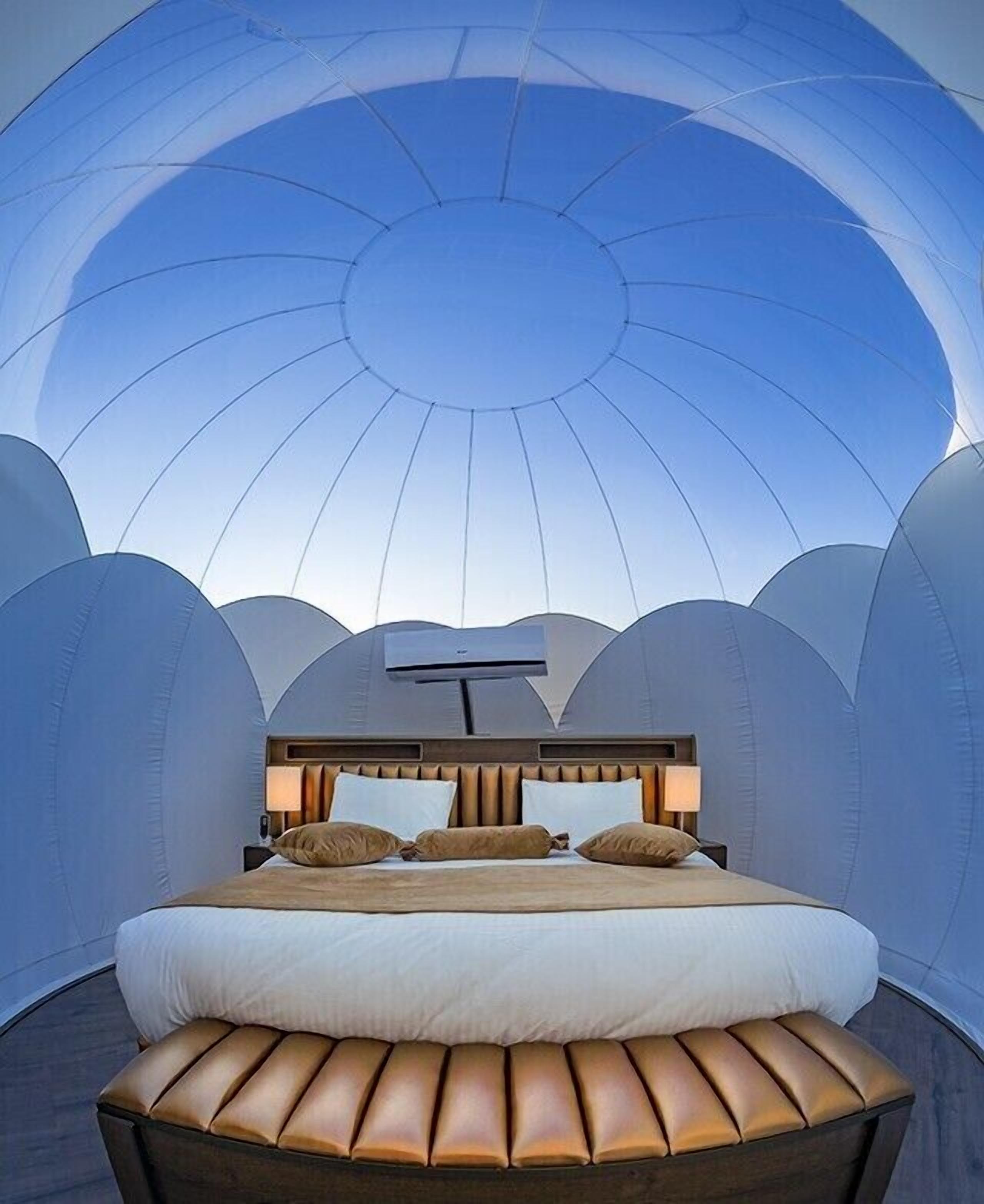 A bedroom with a large bed, a headboard with a light fixture, and a view of the sky through a dome-shaped ceiling.