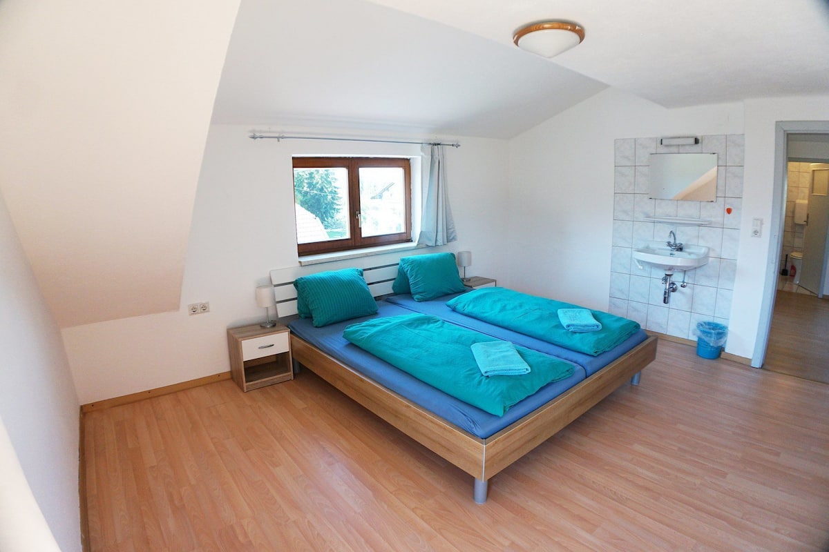 Une chambre à coucher avec un lit en bois, une fenêtre, une table de chevet et une salle de bain équipée d’un lavabo et d’un miroir.