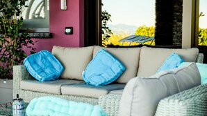 A wicker sofa with blue and light blue cushions, a glass table with a bottle, and a view of greenery and mountains through a window. - B&B Sweet Life (Peschiera del Garda)