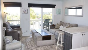 A spacious living area with a sofa, a coffee table, and a dining table. There is a balcony with a view of palm trees and a building.
