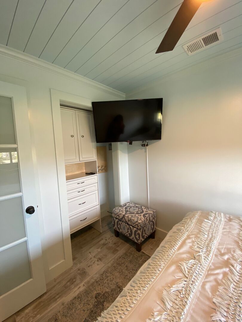 A bedroom with a bed, a TV mounted on the wall, a wardrobe, and a patterned ottoman.