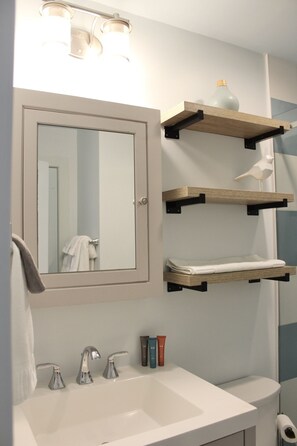 Un baño con lavamanos blanco, dos grifos, un espejo y tres repisas con artículos de aseo.