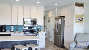 A modern kitchen with white cabinets, a stainless steel refrigerator, a built-in oven, and a microwave. There are two stools and a small table.