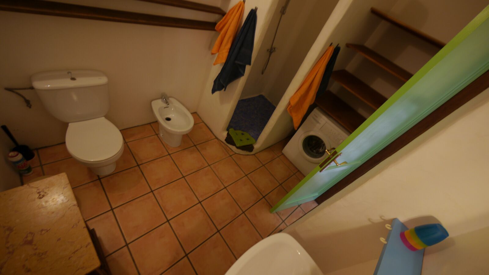 Un baño con inodoro, bidé y lavadora. Hay una mesa de madera y un toallero verde.