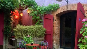 Une terrasse extérieure avec des murs en briques, des volets rouges et une arche en briques. On y trouve deux tables avec des chaises, entourées de plantes en pot et d’une lanterne suspendue.