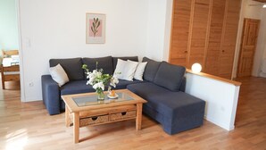 A modern living room with a dark blue sofa, a glass coffee table with a vase of flowers, wooden flooring, and a wooden wardrobe in the background. - Ferienwohnung Krabbe (Kopendorf auf Fehmarn)