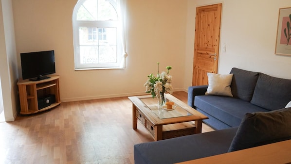 A living room with a TV, a sofa, a coffee table, and a window with curtains. - Ferienwohnung Krabbe (Kopendorf auf Fehmarn)