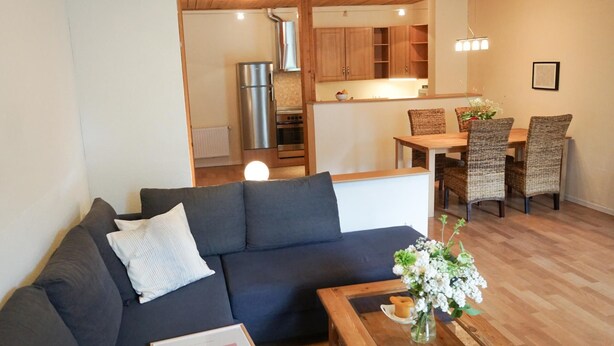 A modern living room with a dark sofa, a coffee table, and a dining area with wicker chairs and a wooden table. - Ferienwohnung Krabbe (Kopendorf auf Fehmarn)