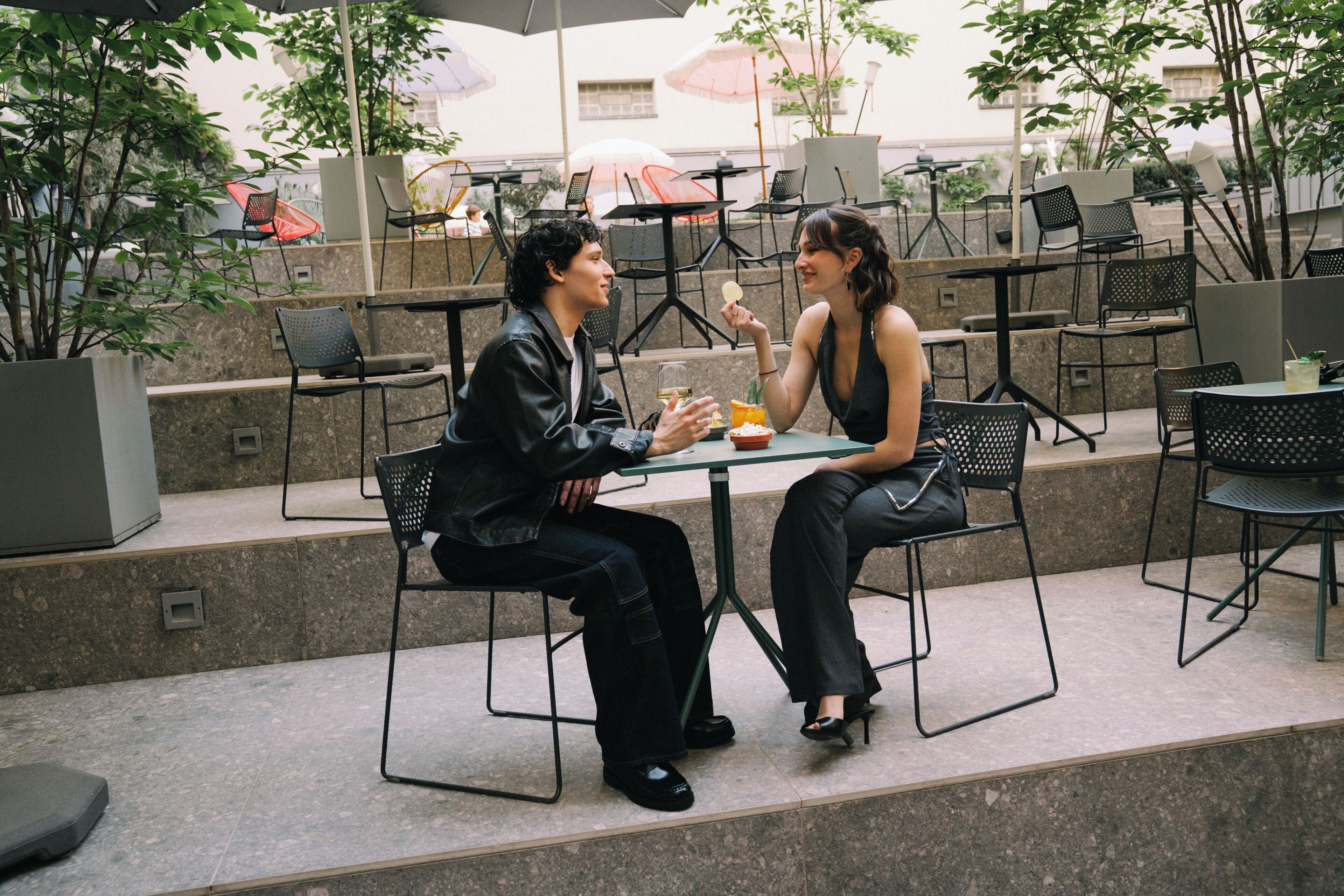 Two people seated at a table with chairs and a glass of drink, outdoors with chairs and umbrellas in the background.