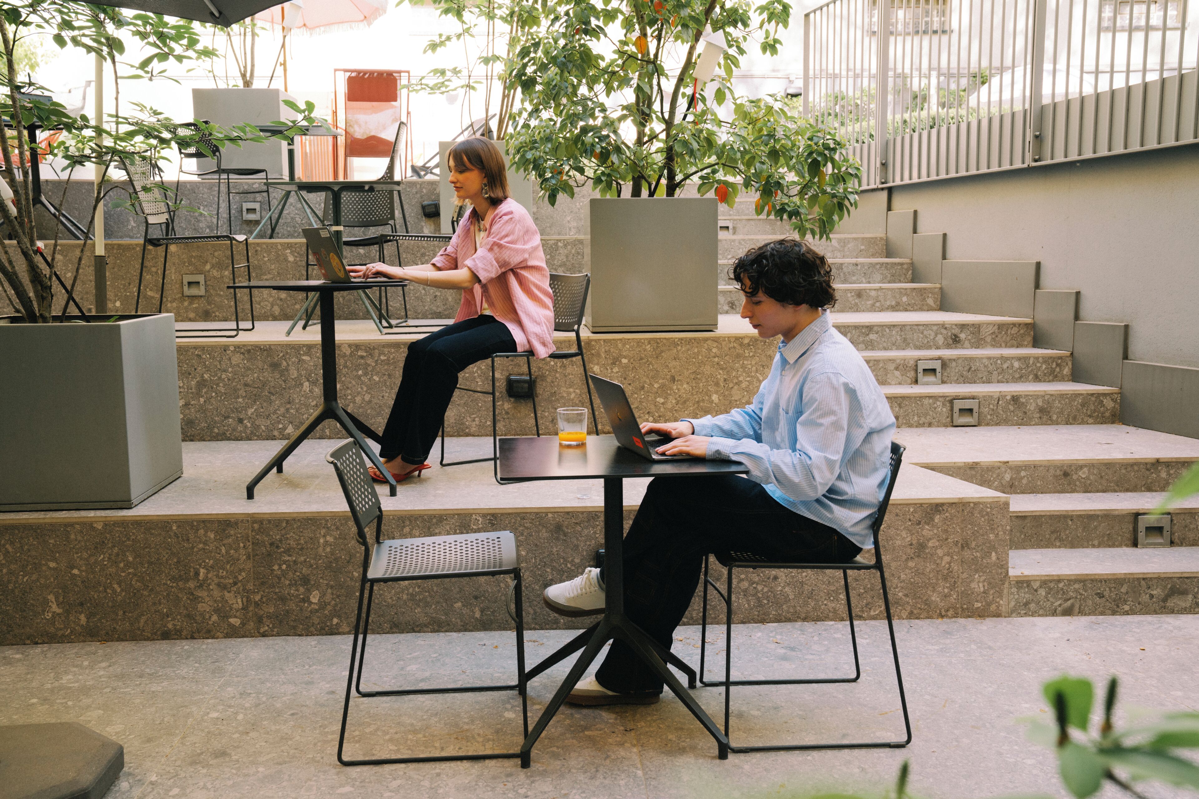 Two people working on laptops at outdoor tables with chairs, surrounded by potted plants and steps.