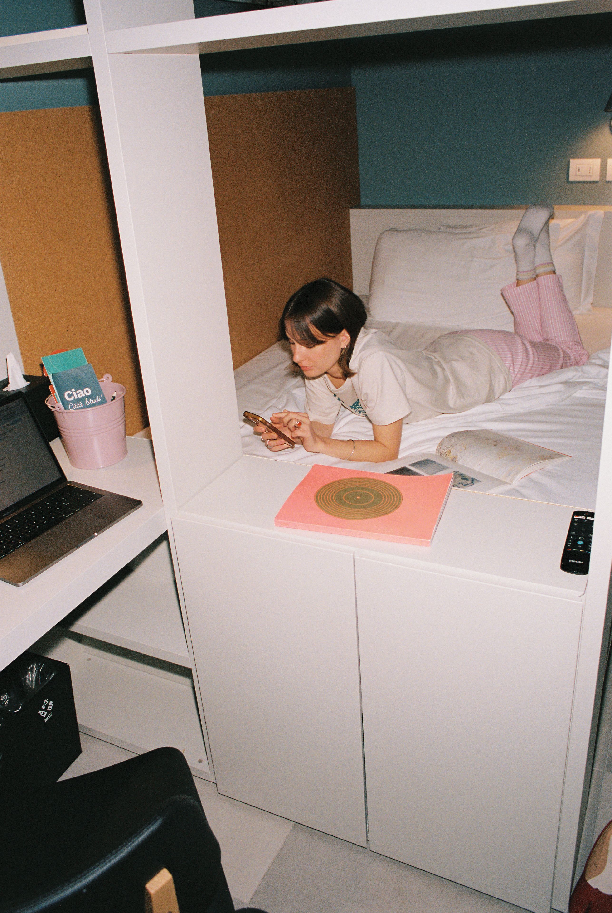Une chambre avec un lit superposé, un bureau avec un ordinateur portable, un livre rose et une télécommande.